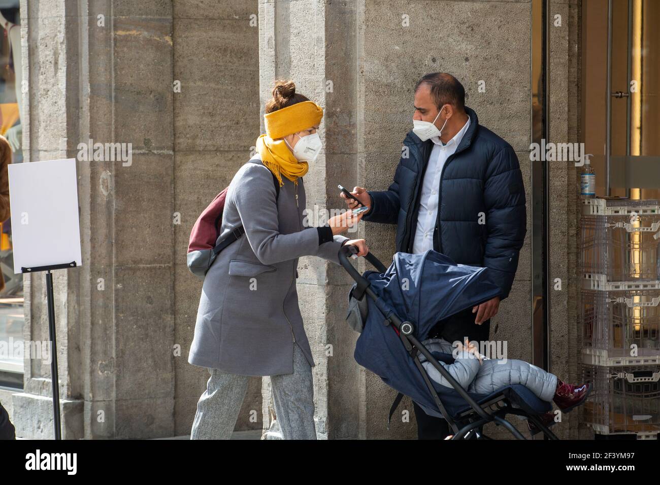 Nachdem eine Mutter einen Termin am Handy vereinbart hatte, darf sie nach Einscannen durch einen Mitarbeiter in das Geschäft. Während die Inzidenz in München weiter ansteigt und evtl. bald die Geschäfte wieder schließen müssen, sind viele Menschen am 18. März 2021 in der Münchner Innenstadt unterwegs und kaufen ein. Die neuen Varianten des Coronavirus betragen in der Landeshauptstadt aktuell fast 70%. * While the incidence is rising and the shops may have to close again, many people on March 18 2021 go to the pedestrian zone in Munich, Germany and buy stuff. The number of new variants in M Stock Photo