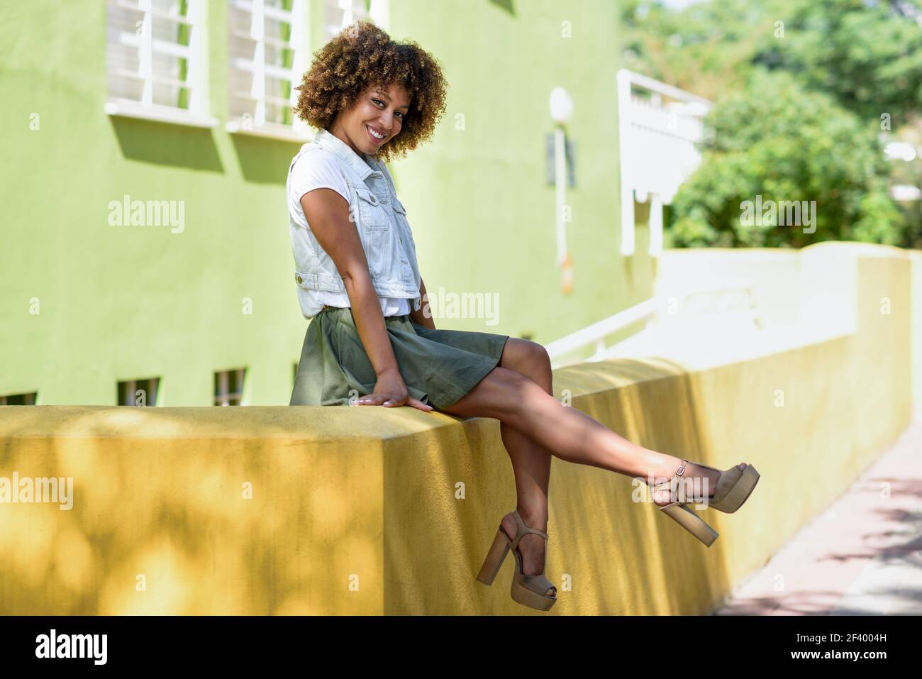 Young black woman, afro hairstyle, sitting on a wall smiling. Girl, model of fashion, wearing casual clothes in urban background. Female with skirt, denim vest and high heels. Stock Photo