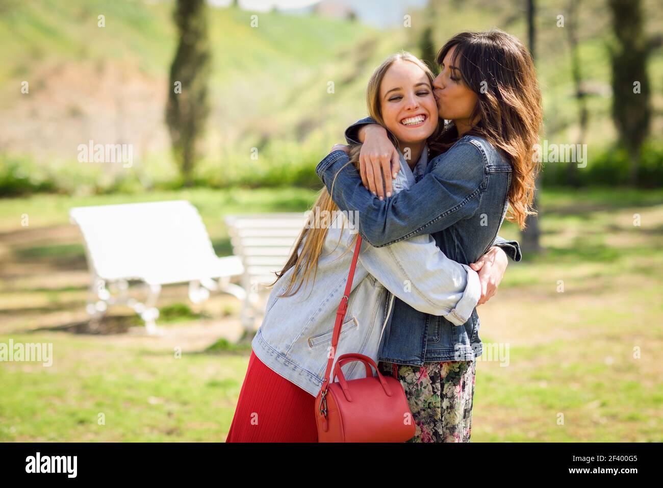 Young woman kissing her friend face outdoors. Blonde and brunette girls wearing casual clothes outdoors. Stock Photo
