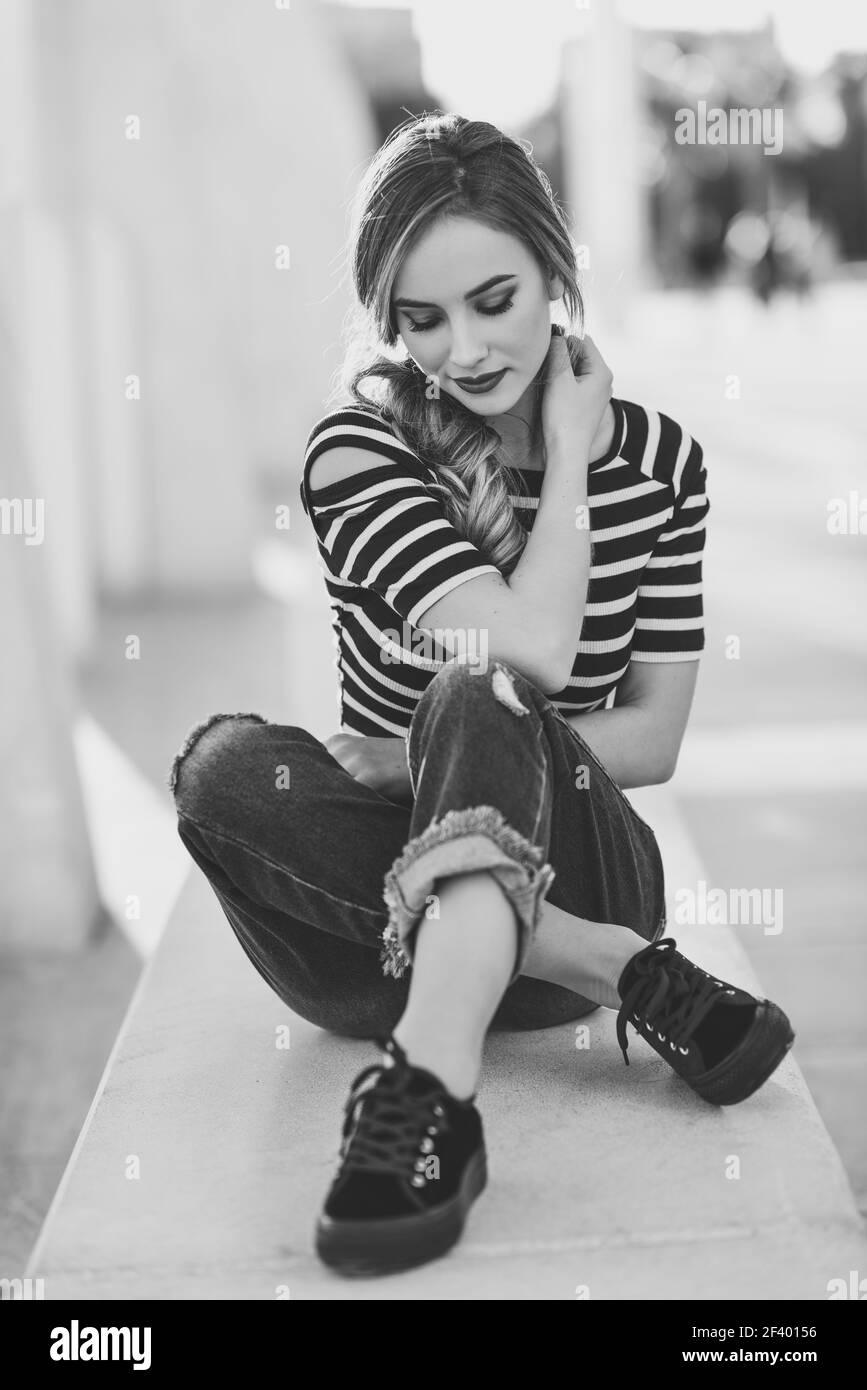 Blonde woman, model of fashion, sitting in urban background.. Blonde woman, model of fashion, sitting on a bench in urban background with eyes closed. Thoughtful young girl wearing striped t-shirt and blue jeans in the street. Pretty russian female with pigtail. Stock Photo