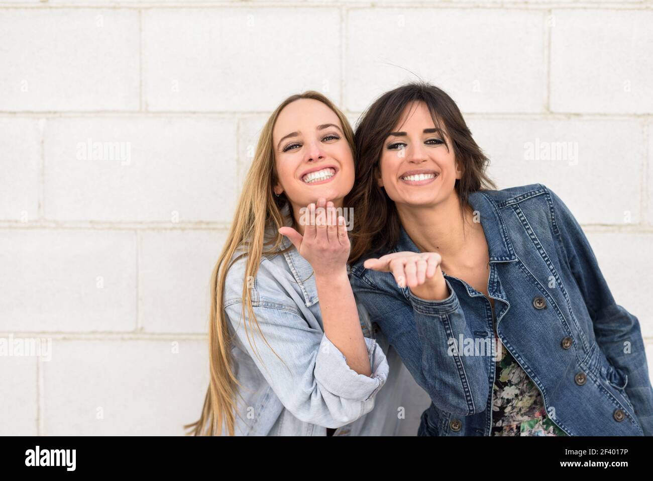 Two young women blowing a kiss on urban wall.. Two beautiful girls blowing a kiss on urban wall outdoors. Young women wearing casual clothes. Stock Photo