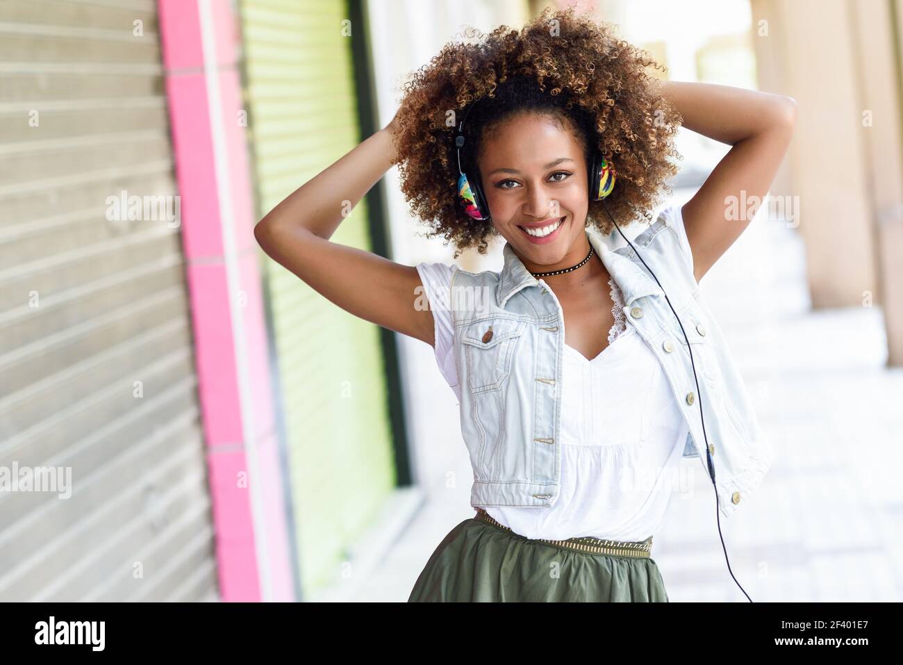 Young black woman, afro hairstyle, in urban street with headphon. Young attractive black woman in urban street listening to the music with headphones. Girl wearing casual clothes with afro hairstyle Stock Photo
