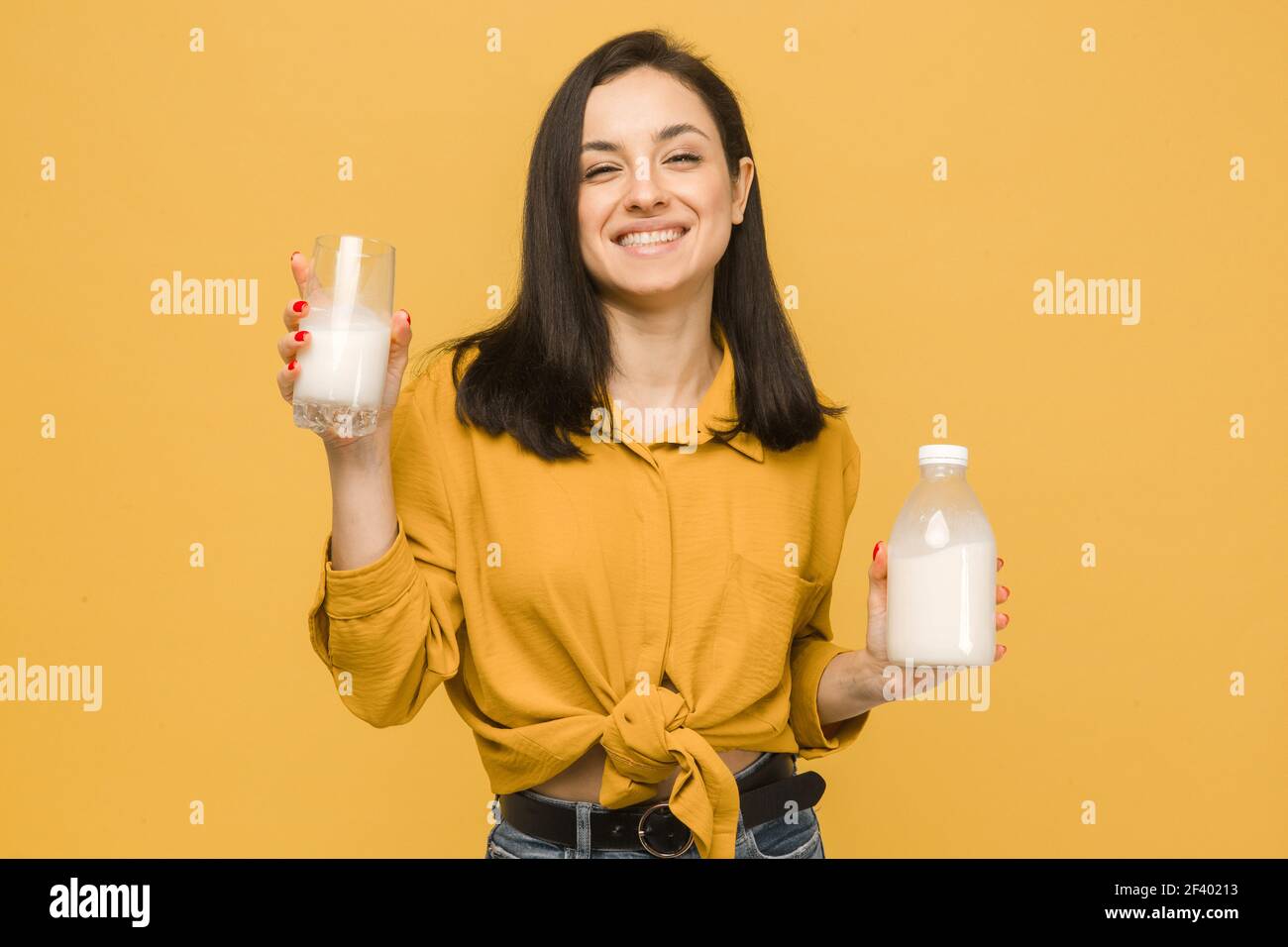 Concept photo of young female is holding glass and bootle of milk. Wears yellow shirt, isolated yellow color background Stock Photo