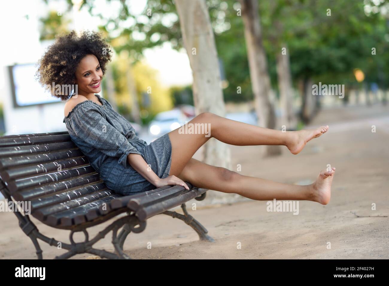 Young black woman with afro hairstyle smiling in urban backgroun. Young black woman with afro hairstyle sitting on a bench in urban background moving her legs. Mixed girl wearing casual clothes. Stock Photo