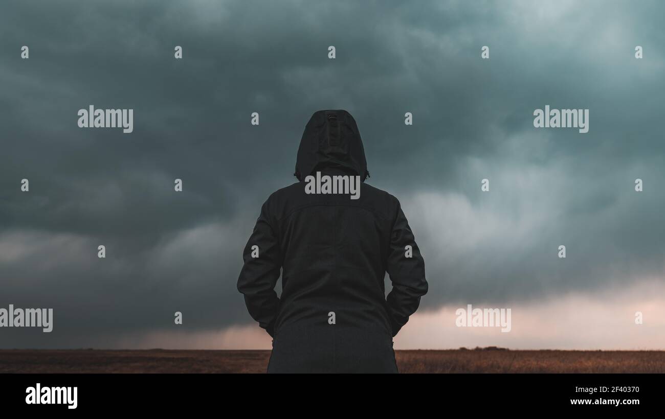 Rear view of female person wearing hooded jacket against dark moody dramatic clouds at sky, woman looking into uncertain ominous future Stock Photo