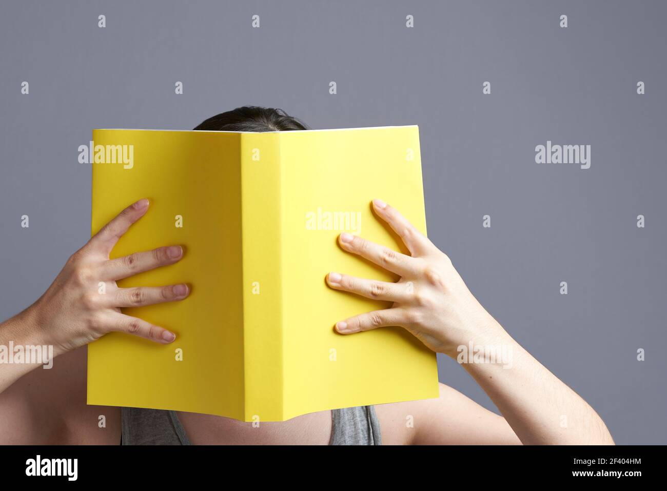 Young woman with her face hidden behind a yellow book that she holds with both hands, open, reading it. Portrait with gray background and copy space. Stock Photo