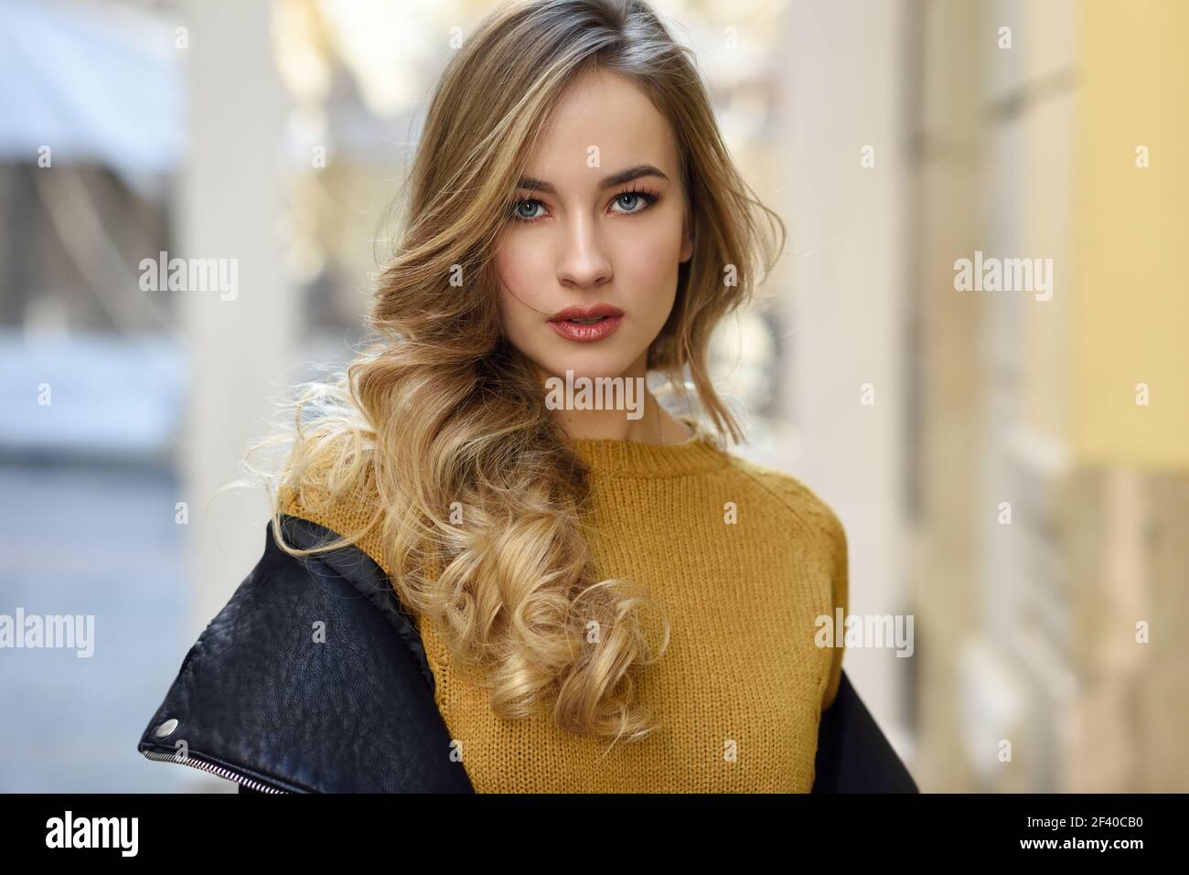 Blonde woman in urban background. Beautiful young girl wearing black leather jacket and mini skirt standing in the street. Pretty russian female with long wavy hair hairstyle and blue eyes. Stock Photo