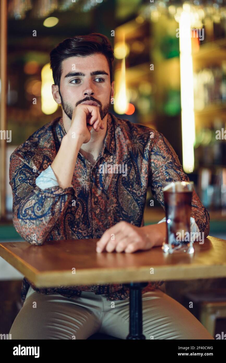 Thoughtful bearded man wearing casual clothes sitting in a modern pub. Guy with beard and modern hairstyle drinking a cola. Stock Photo