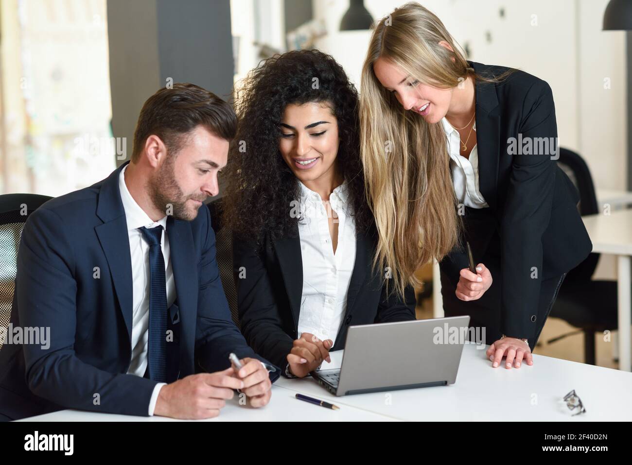 Multi-ethnic group of three businesspeople meeting in a modern office. Two women and a man wearing suit looking at a laptop computer. Stock Photo
