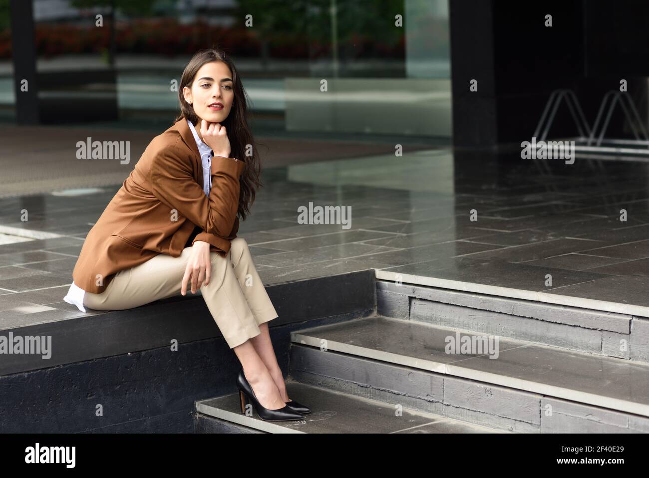 Young businesswoman sitting outside of office building. Beautiful woman wearing formal wear. Young girl with brown jacket and trousers in urban background. Stock Photo