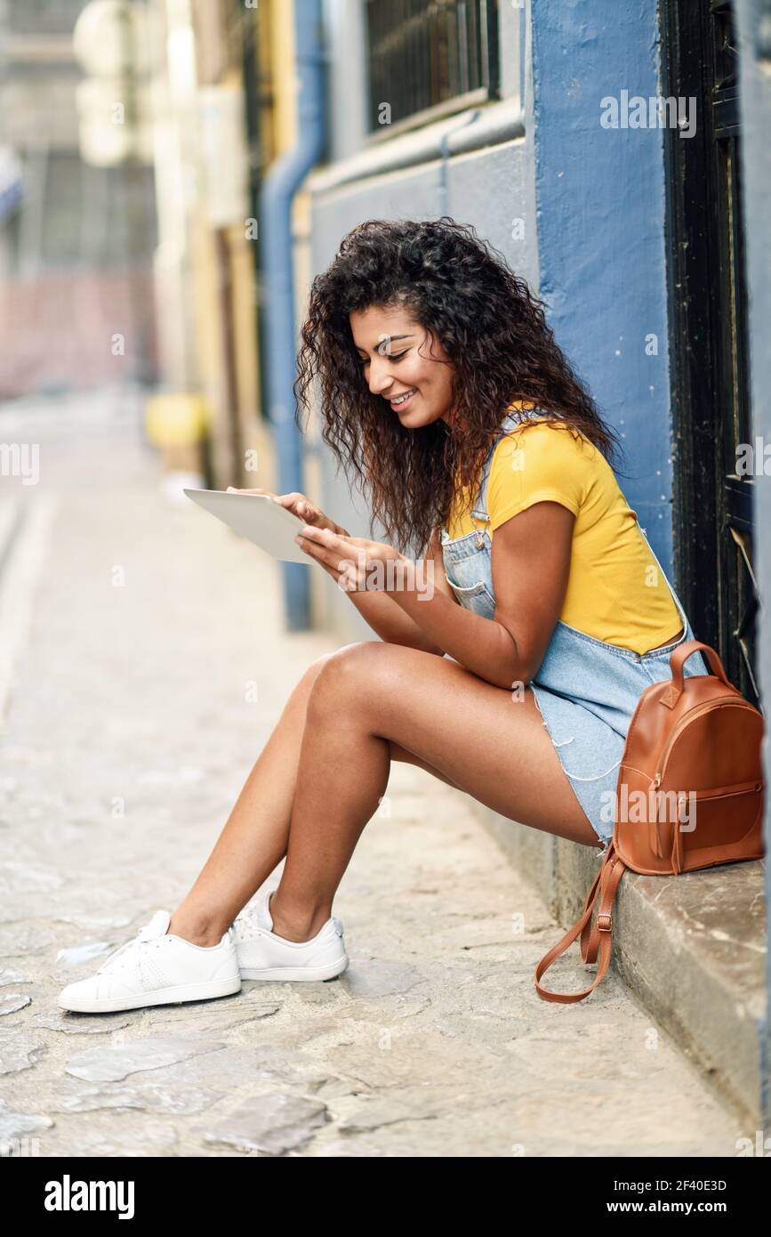 Young Arab woman looking at her digital tablet outdoors. African girl wearing casual clothes inurban background. Curly hairstyle. Stock Photo