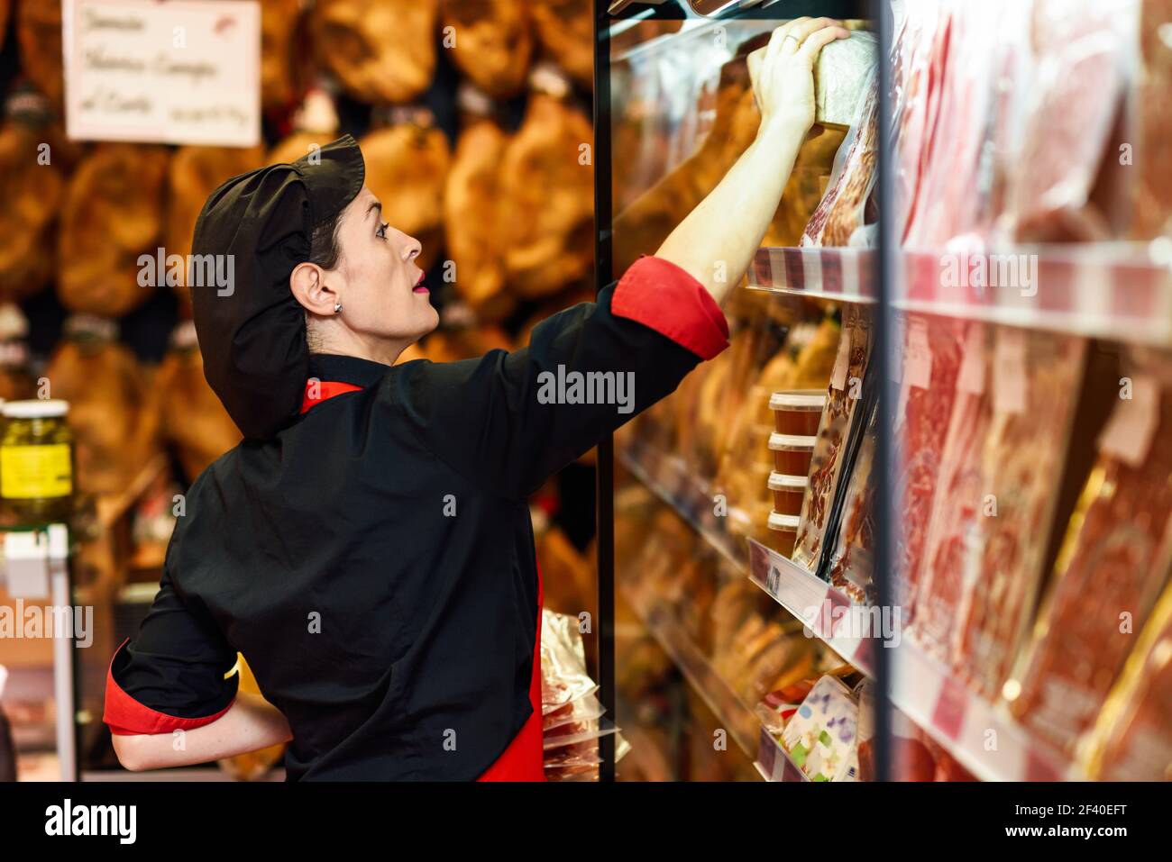 Portrait of female worker taking products in butcher shop. Refrigerated display case for sausage packages, ham and cheese Stock Photo