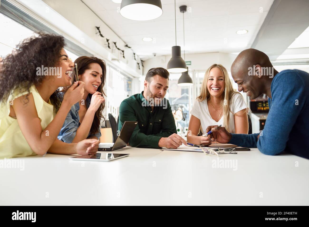 Five young people studying with laptop and tablet computers on white desk. Beautiful girls and guys working toghether wearing casual clothes. Multi-ethnic group smiling. Stock Photo