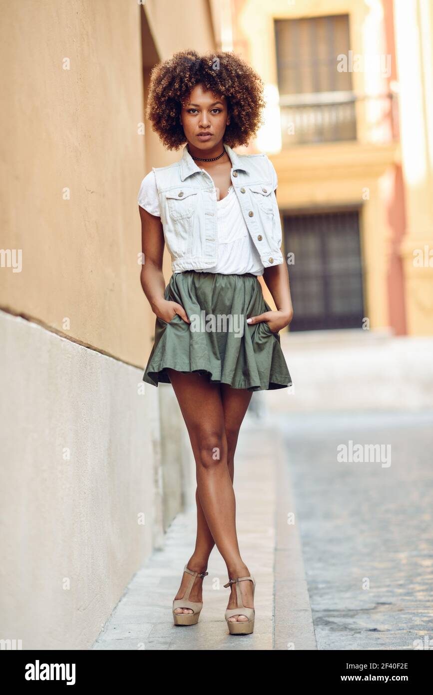 Young black woman, afro hairstyle, standing in the street. Girl wearing casual clothes in urban background. Female with skirt, denim vest and high heels. Stock Photo