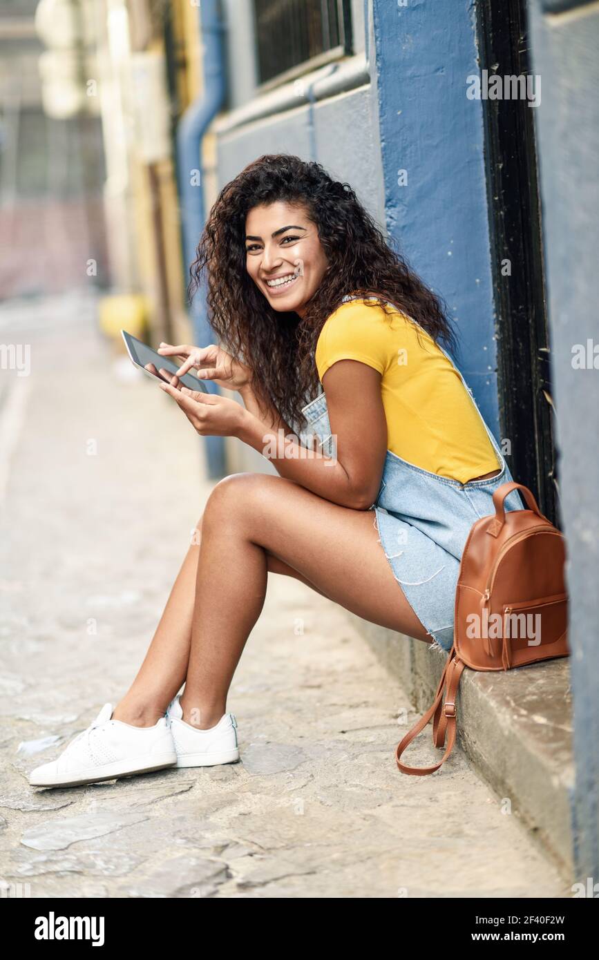 Happy Arab woman sitting on urban step with a digital tablet. African girl wearing casual clothes. Young traveler female with curly hairstyle smiling. Stock Photo