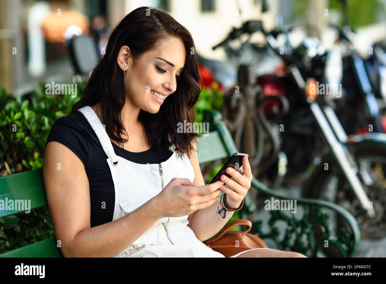 Smiling young woman looking at smart phone outdoors. Girl wearing white denim dress sitting in urban bench. Technology concept. Stock Photo