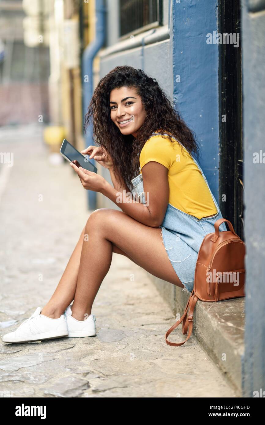 Happy Arab woman sitting on urban step with a digital tablet. African girl wearing casual clothes. Young traveler female with curly hairstyle smiling.. Happy Arab woman sitting on urban step with a digital tablet Stock Photo