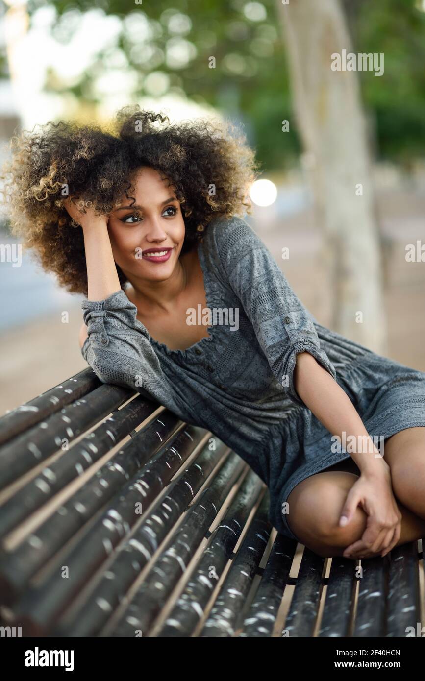 Funny black woman with afro hairstyle sitting on a bench in urban background. Mixed girl wearing casual clothes. Young black woman with afro hairstyle smiling in urban background Stock Photo