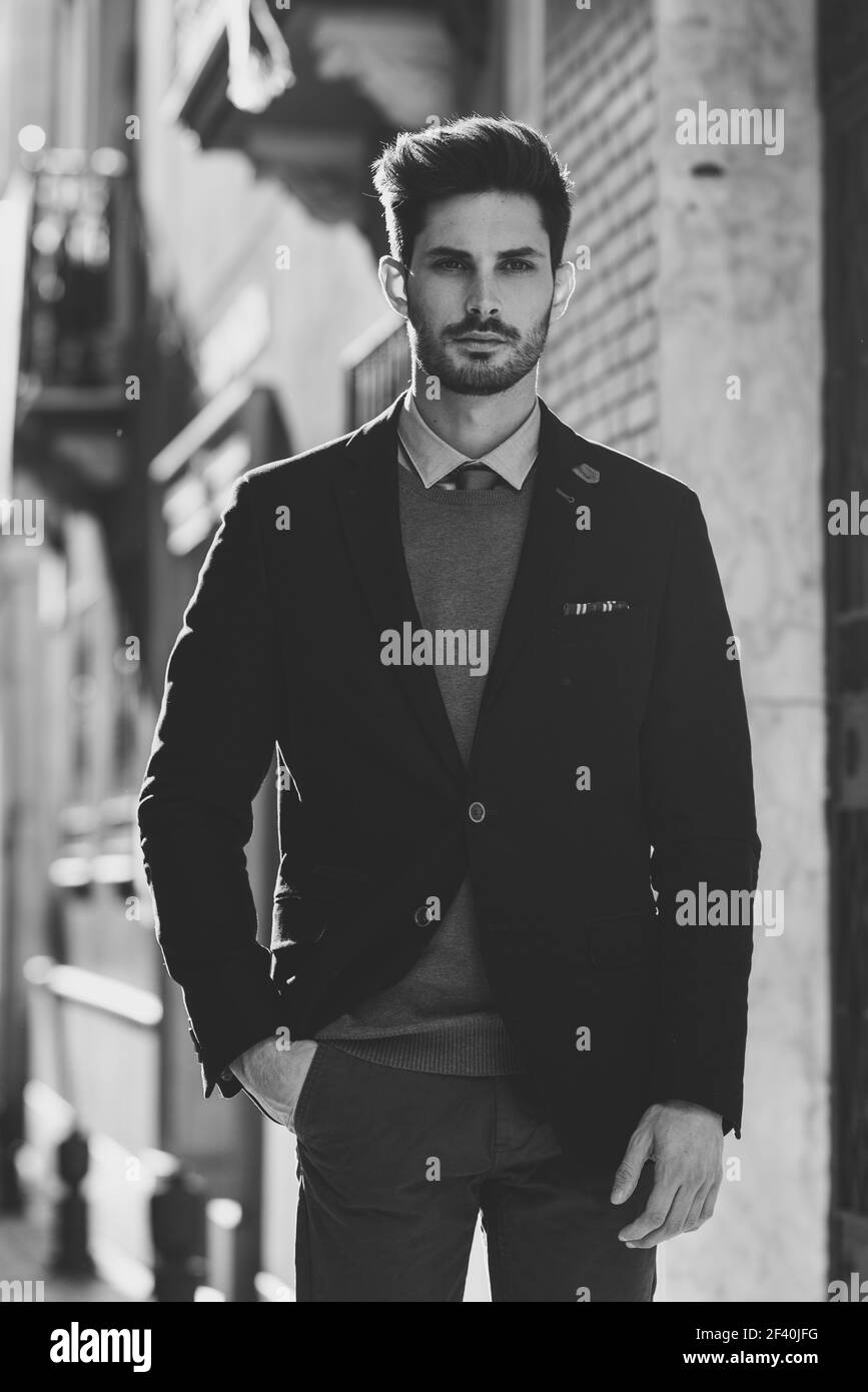 Attractive man in the street wearing british elegant suit. Young bearded businessman with modern hairstyle in urban background. Black and white photograph.. Attractive man wearing british elegant suit in the street. Modern hairstyle. Stock Photo