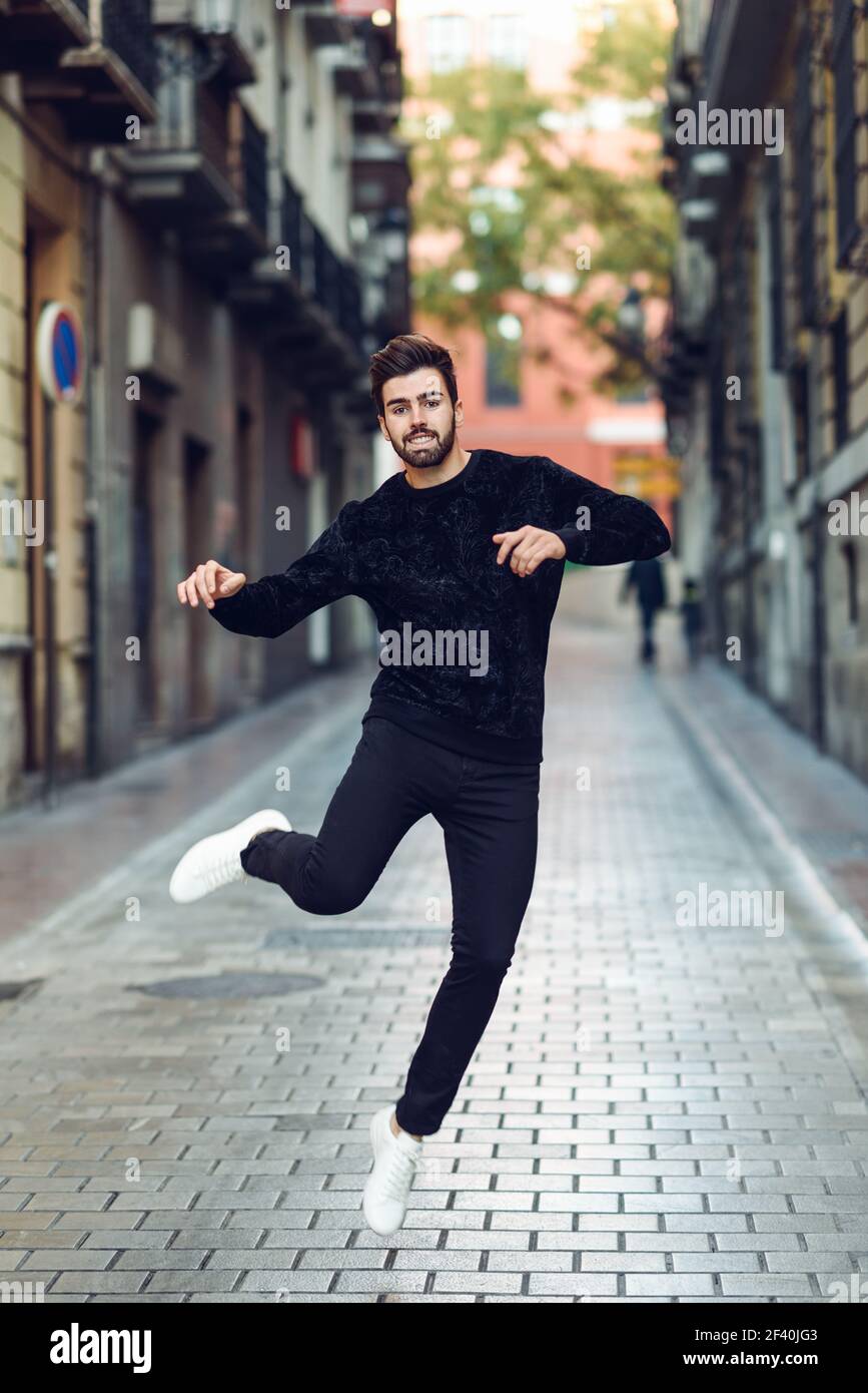 Young bearded man jumping in urban background with open arms wearing casual clothes. Guy with beard and modern hairstyle in the street.. Young bearded man jumping in urban background with open arms wearing casual clothes. Stock Photo