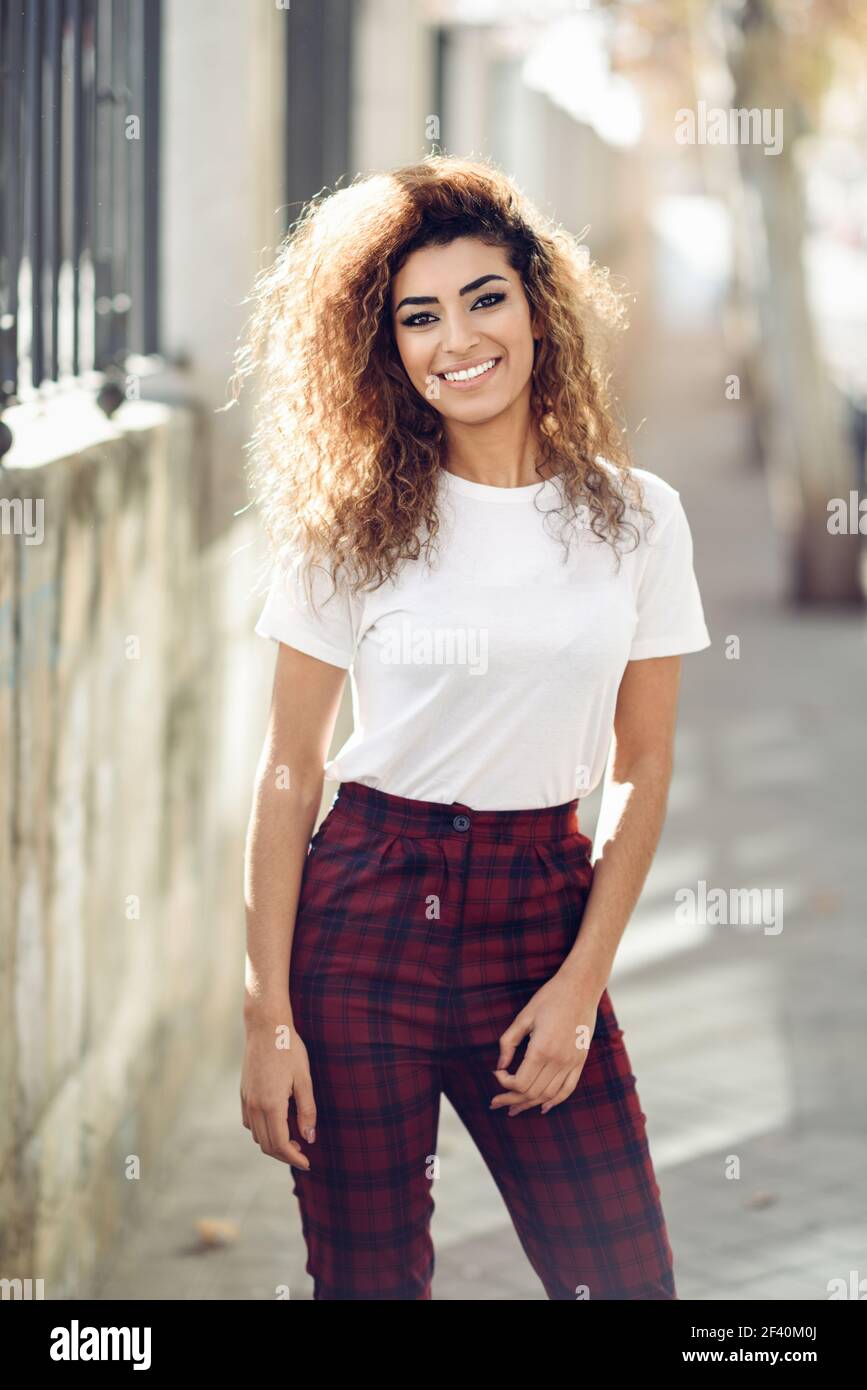 Beautiful young arabic woman with black curly hairstyle. Arab girl in  casual clothes in the street. Happy female wearing white t-shirt and  checked pants.. Arab girl in casual clothes in the street