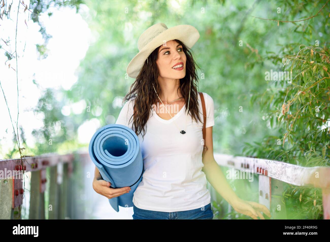 Beautiful hiker young woman with backpack, with blue eyes, wearing straw hat, hiking in the countryside with a blue mat in her hand.. Woman with backpack standing on rural bridge. Stock Photo