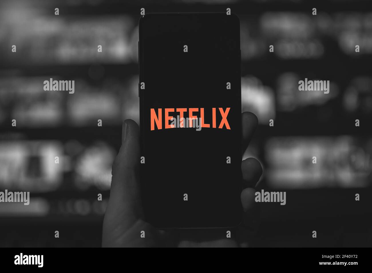 Man holding a smartphone with red Netflix logo with black and white blurred TV background. Selective color. Popular stre Stock Photo