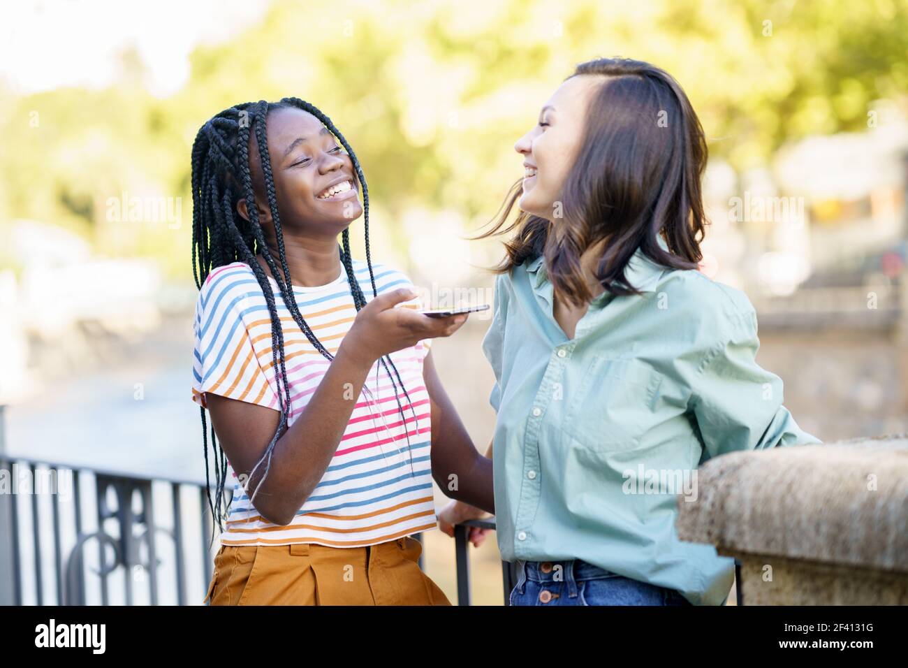 Two female friends recording a voice note with smartphone outdoors. Multiethnic friends.. Two female friends having fun together on the street. Multiethnic friends. Stock Photo