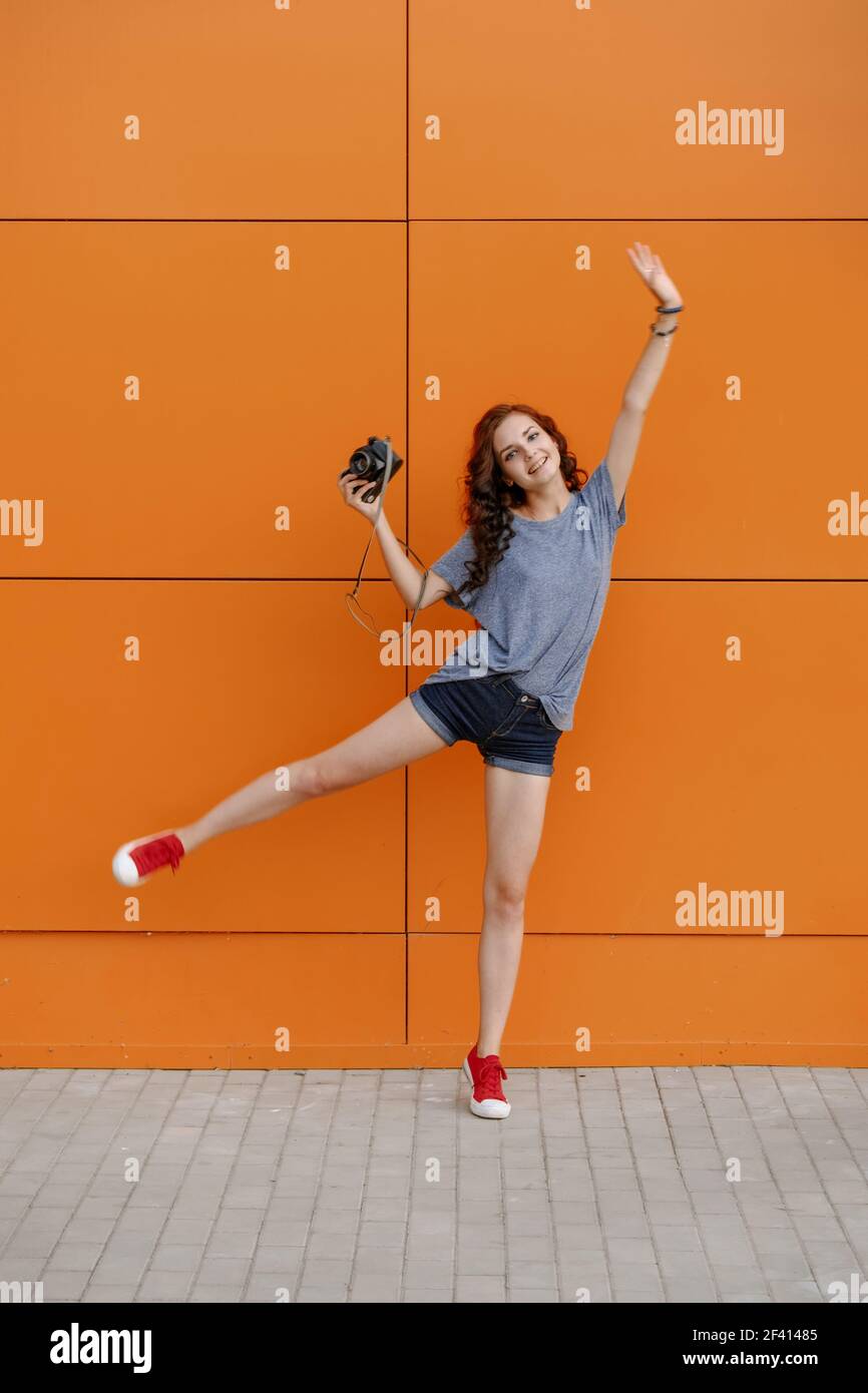 Hipster girl with vintage photo camera in one hand standing on one leg in front of orange wall, copy space, toned image. Hipster girl with vintage photo camera in one hand standing on one leg, copy space, toned image Stock Photo