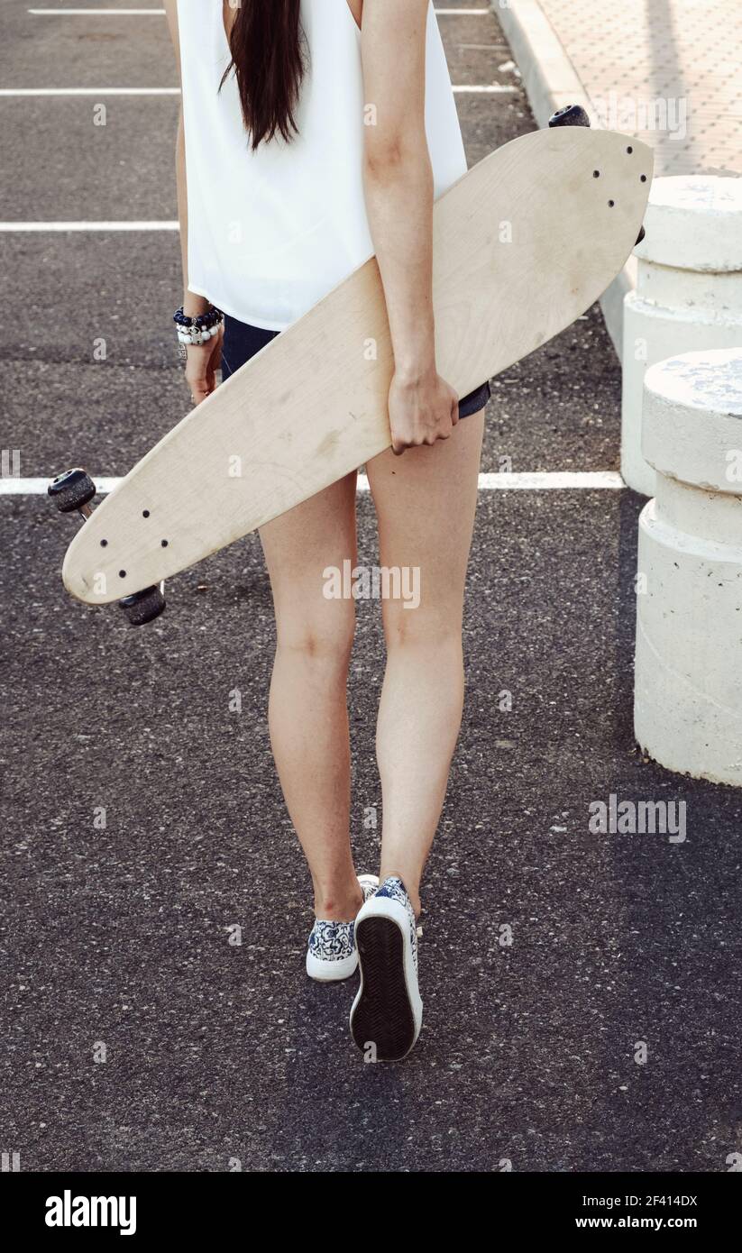 Rear view of skater girl walking away holding her longboard behind her ...
