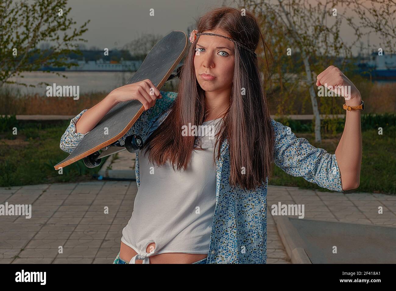 Funny girl in boho attire with skatebord on her shoulder make I-am-powerful gesture by her left hand. Funny girl in boho attire with skatebord on her shoulder make I-am-powerful gesture Stock Photo