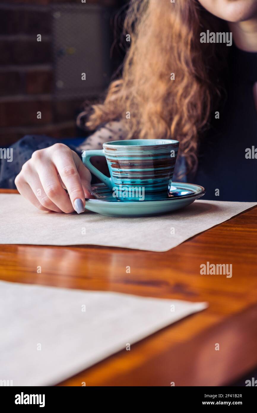 The red-haired girl sits alone at a table in a cafe, the focus on the cup facing her. place for inscriptions in the foreground. On the table are two sheets of craft paper.. Red-haired girl sits alone at a table in a cafe, focus on the cup Stock Photo
