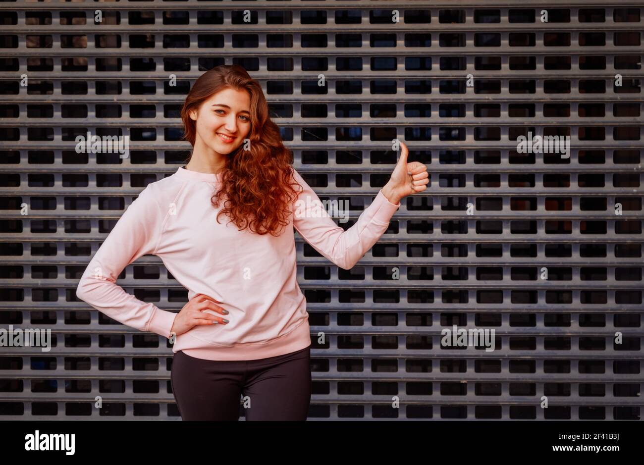 Happy redhead girl shows us thumb up sign in front of metal grid. Happy redhead girl shows us thumb up sign Stock Photo
