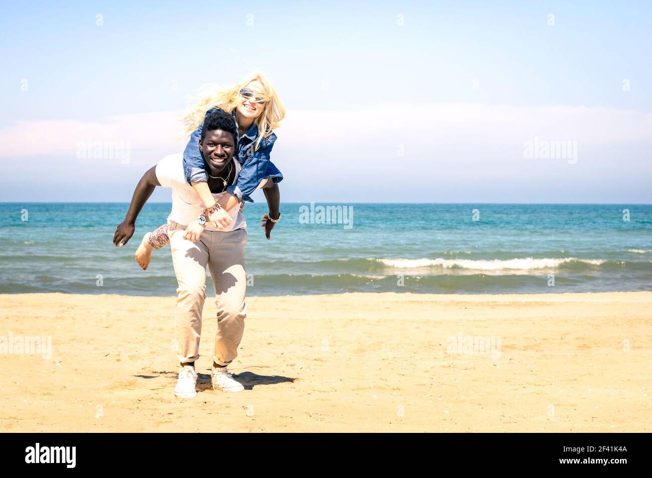Young multiracial couple at the beach having fun with piggyback jump - Happy mixed race boyfriend and girlfriend playing Stock Photo