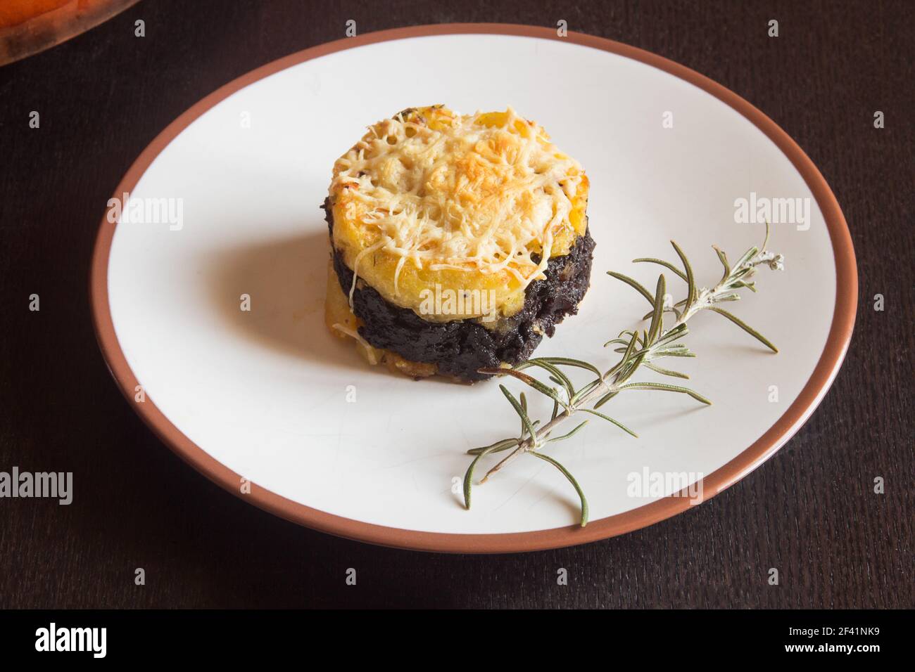 Presentation food plate of potato cake with black pudding cream, grated cheese, and a fresh branch of rosemary on a wooden table. Homemade recipes. Stock Photo