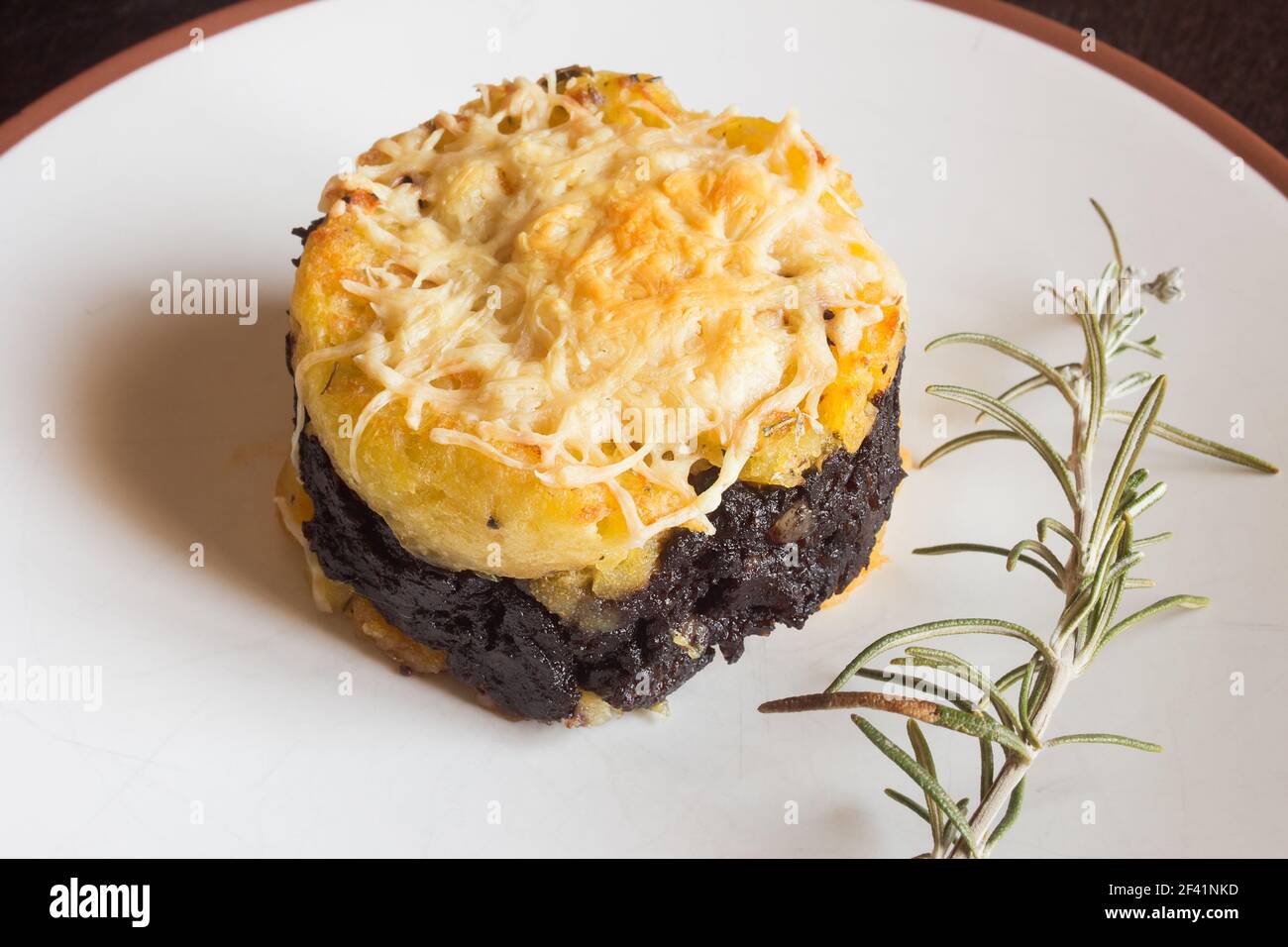 Close-up of a buttered potato pie with blood sausage topped with melted grated cheese on a plate next to a fresh sprig of rosemary. Homemade recipes. Stock Photo