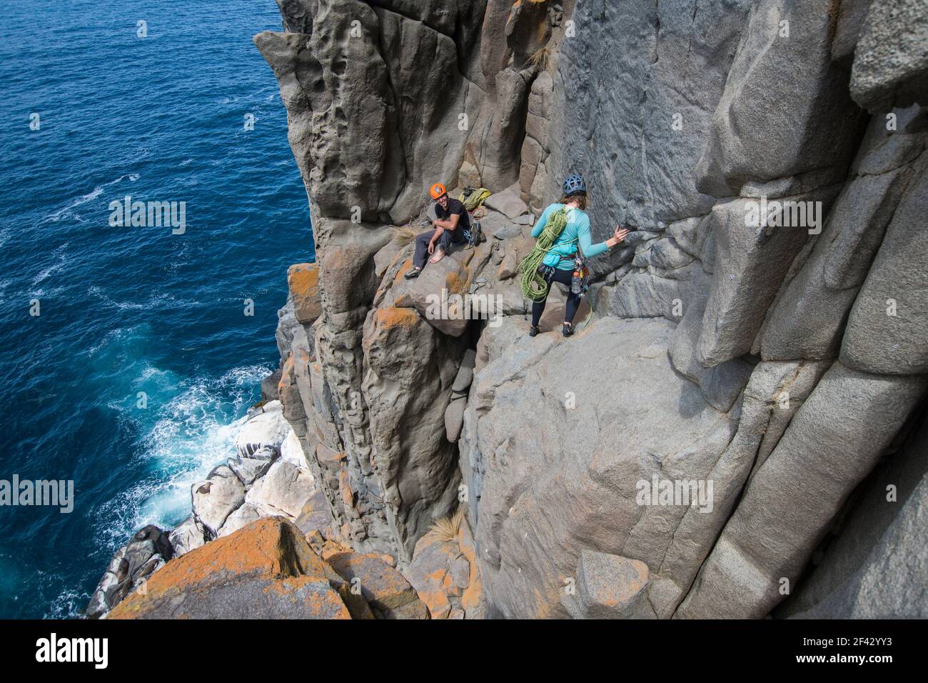 A couple of intrepid rockclimbers explore the featured sea cliffs of Cape Raoul, smiling at each other as they traverse the exposed terrain above a seal colony, in Tasmania, Australia. Stock Photo