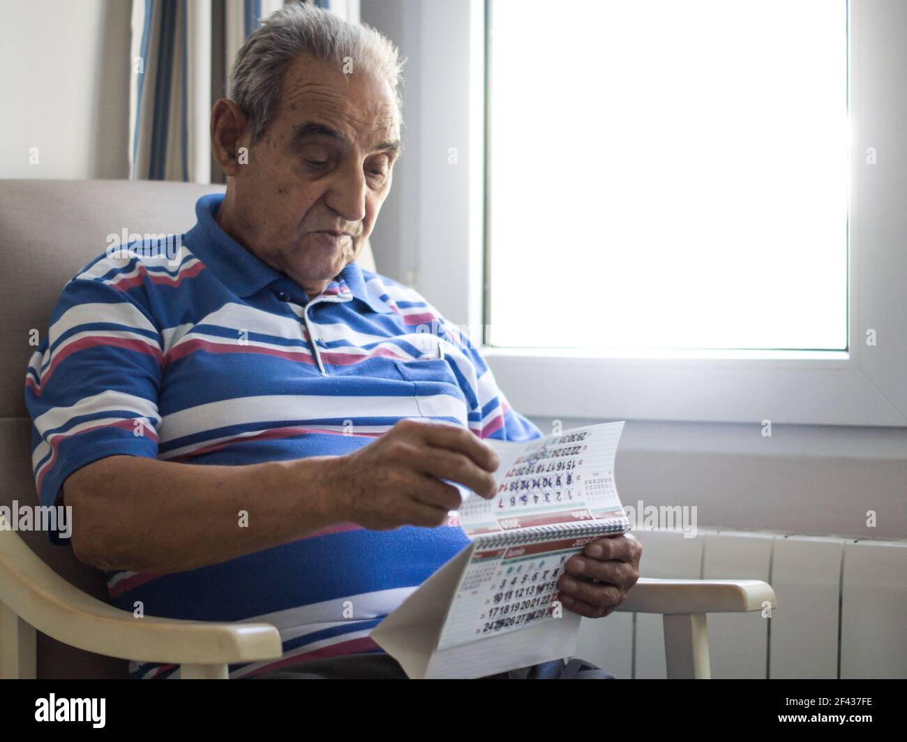 Elderly person looking at a calendar sitting in an armchair next to a very bright window with a striped shirt Stock Photo