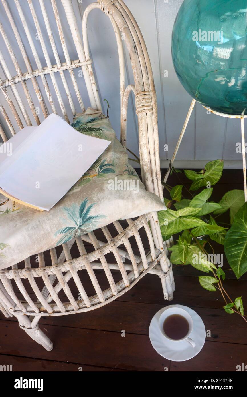 Retro white cane chair with a cushion and book, a cup of tea, plants and ornaments against wall and a wooden floor in casual tropical setting. Stock Photo