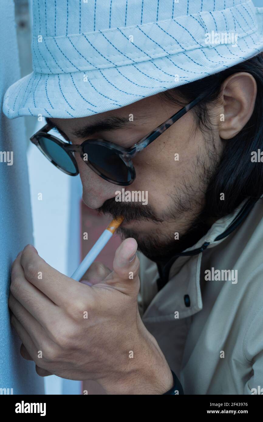man with long hair smoking a cigarette. hipster smoking cigarette. man with  hat and sunglasses trying to light a cigarette with a lighter. May 31 worl  Stock Photo