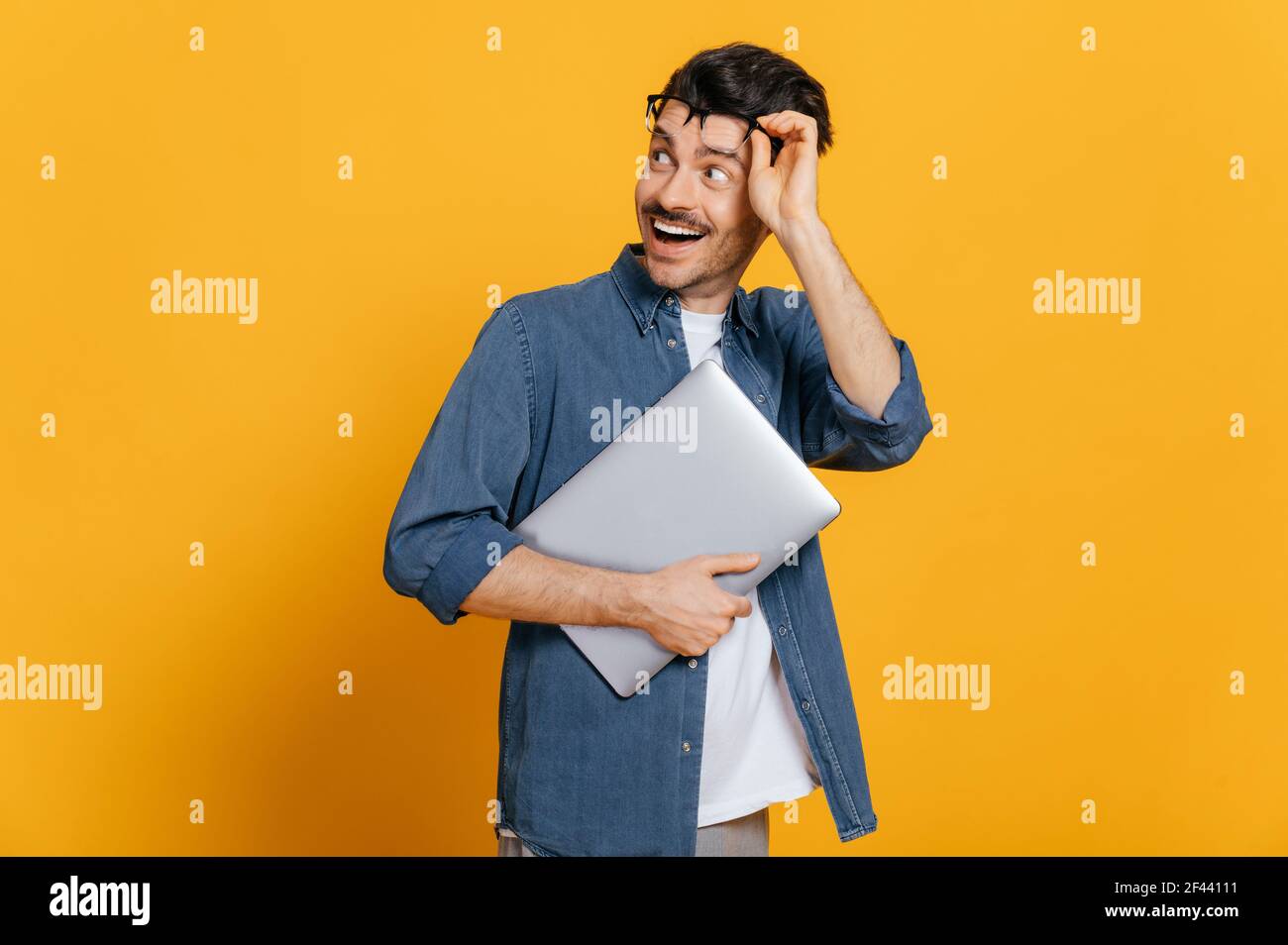 Amazed excited handsome stylish smart guy in glasses and in denim shirt, holds a laptop at hand, looks surprised towards empty space taking off glasses, smiling, isolated orange background, copy space Stock Photo