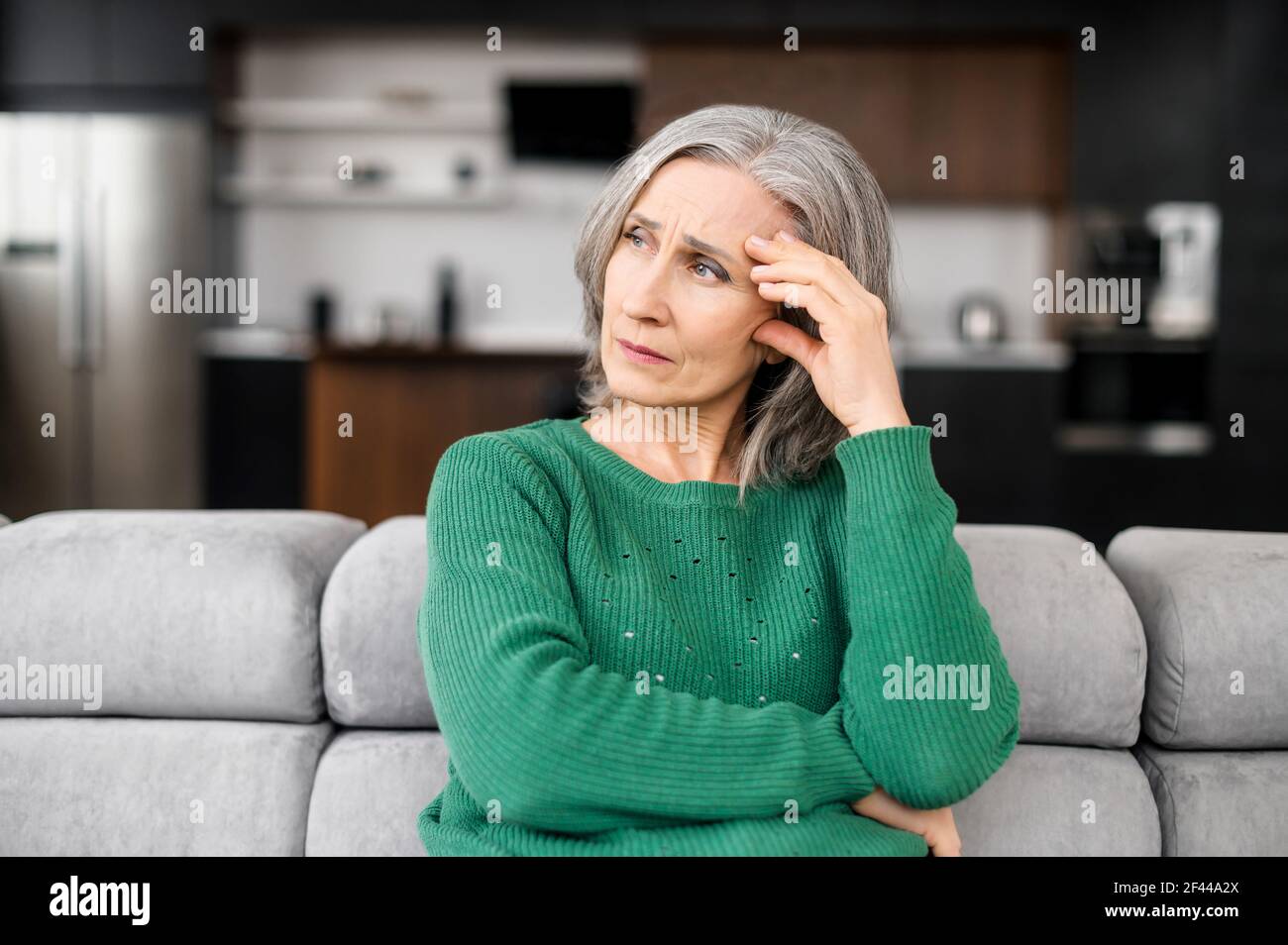 Upset depressed senior gray-haired lady wearing green jumper touching temple, sitting on sofa, looking in the window, feeling worried and stressed about the future and thinking about own problems Stock Photo