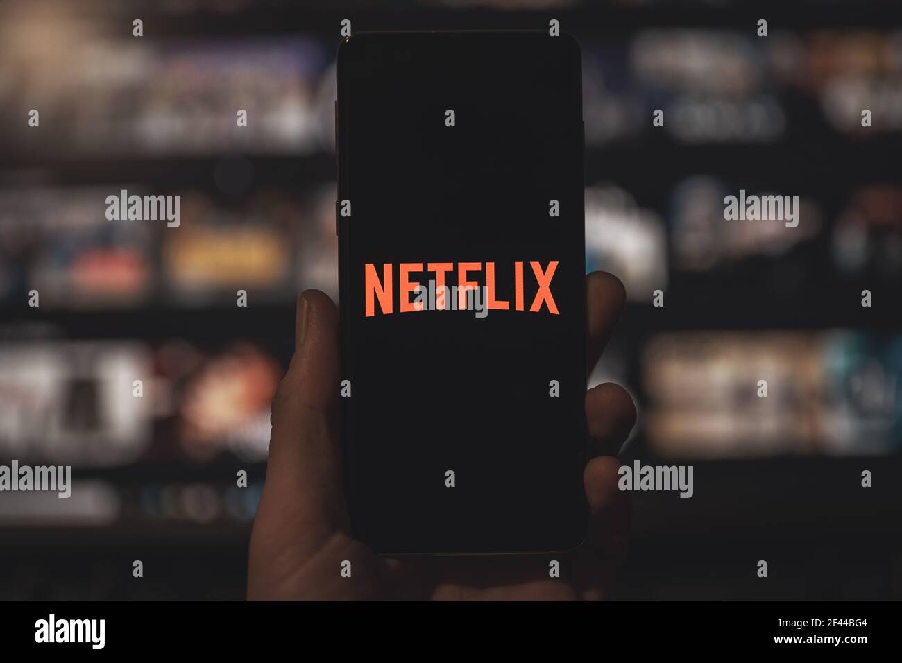 Man holding a smartphone with red Netflix application logo with blurred TV background. Popular streaming service. Stock Photo