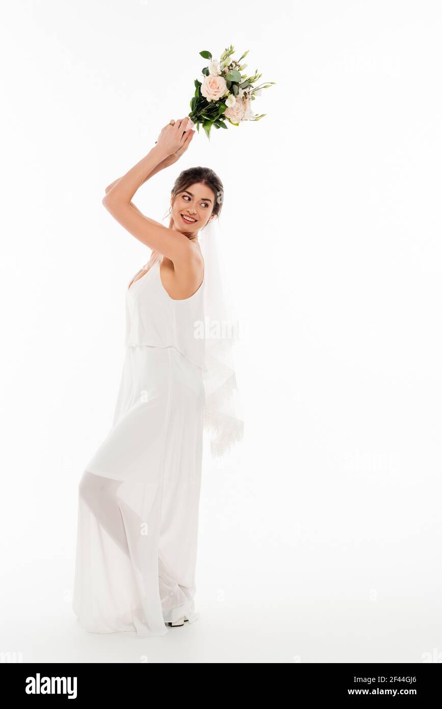 full length view of cheerful bride throwing wedding bouquet on white