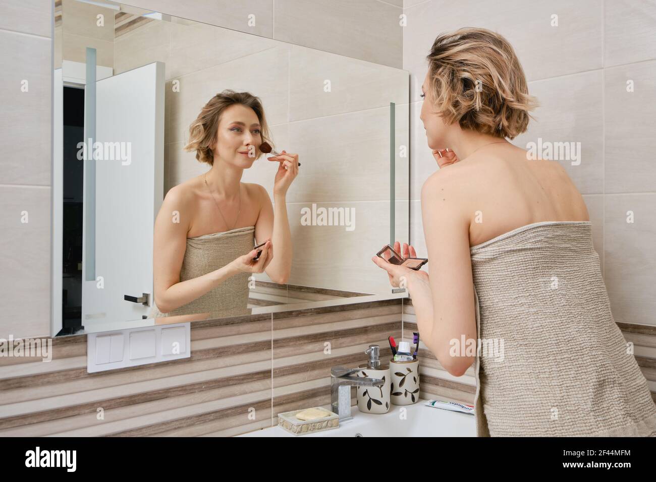 Woman applying blush on face while standing in front of the mirror in the b...