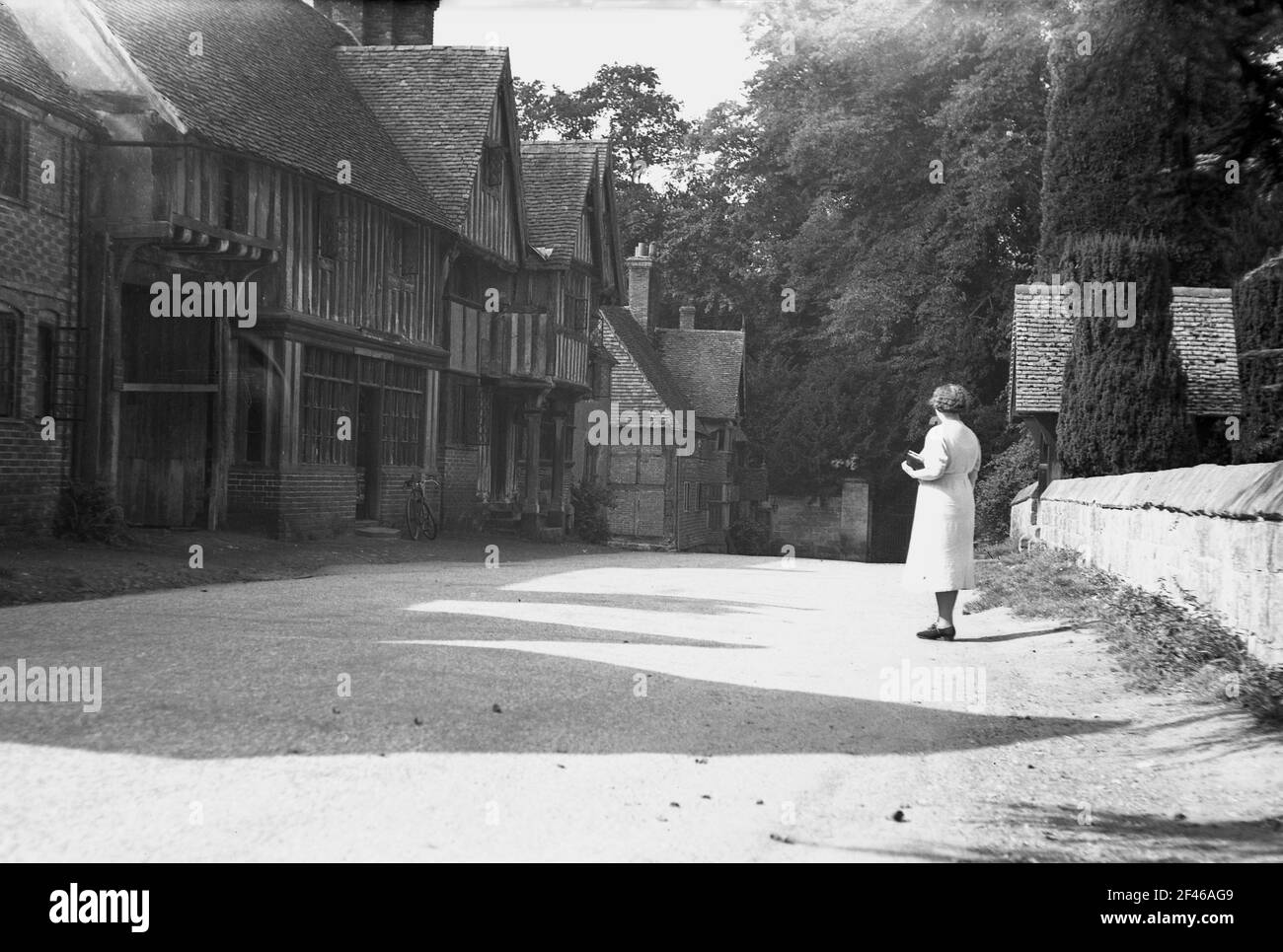 1950s, historical, a lady standing on a gravel road at the small village of Chiddingstone, Kent, England, UK, looking at distinctive wooden-framed Tudor buildings. The old tavern, the Castle Inn is at the end of the street at the entrance to the Castle itself. The old buildings in the village were taken over by the National Trust in 1939. Stock Photo