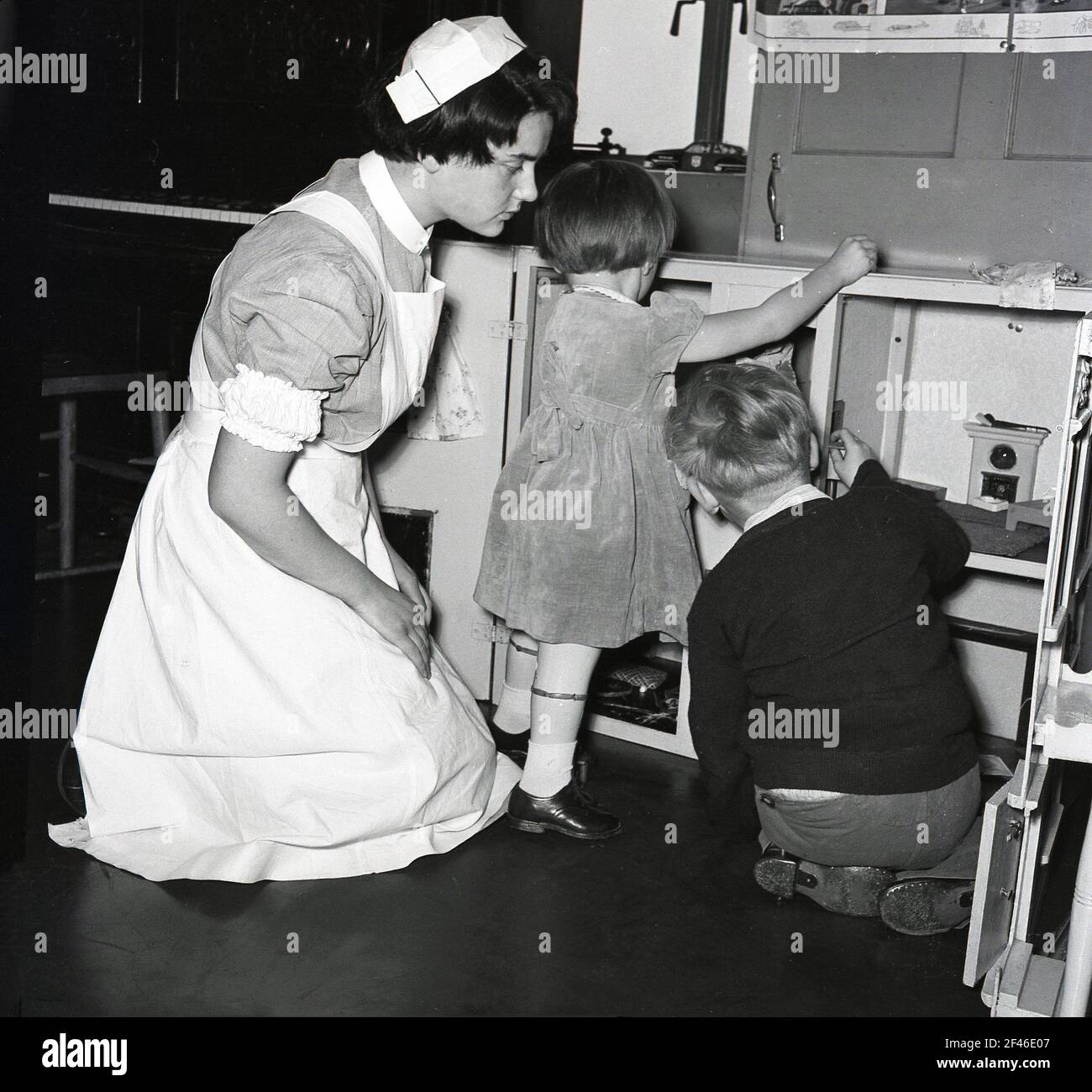 1950s, historical, a young female nurse supporting a little girl and boy as they play at a cupboard with toys in a ward in a children's hospital, England, UK. Stock Photo