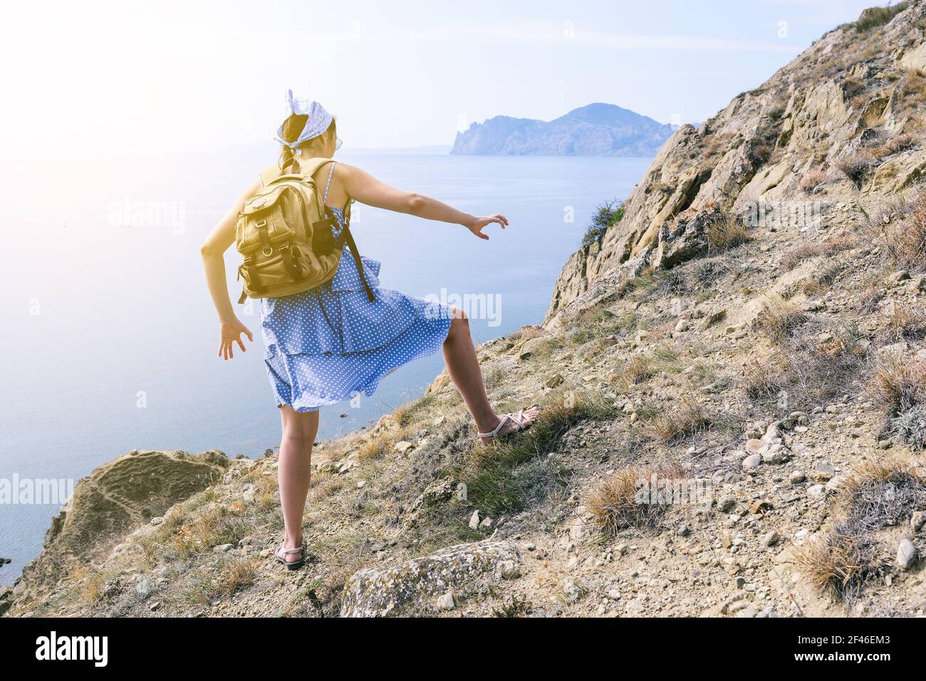 Girl in a blue dress and with a backpack climbs the mountain. Hiker on top of a mountain with a beautiful panorama of the sea. Mountain hiking and spo Stock Photo