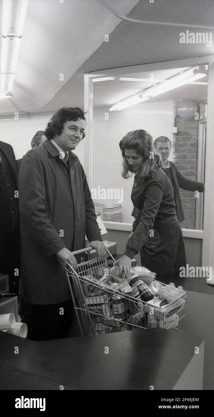 1970s, historical, man in a raincoat  in the pay area of new self-service store or supermarket with a loaded shopping trolley being held in the queue by a female employee, London, England UK. Stock Photo