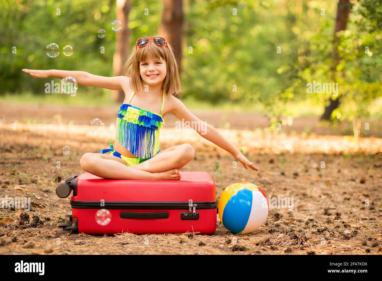 Little child girl wear swimsuit sitting on suitcase in summer forest with hands up, show plane. Dream journey. Vacation concept Stock Photo